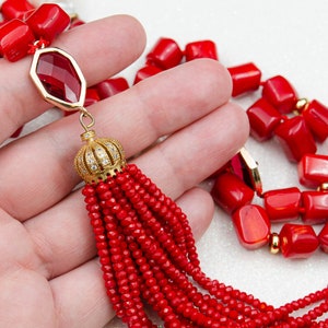 May include: A necklace featuring vibrant red chunky beads and faceted red stones set in gold-toned metal frames. The design includes a prominent gold-toned crown-shaped cap adorned with small clear crystals, from which a long red beaded tassel hangs.
