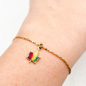 Puede incluir: Una pulsera dorada con un dije de hoja de arce colorido. El dije presenta esmalte rojo, amarillo y verde. La pulsera se lleva en la muñeca.