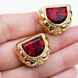 May include: Two gold-toned earrings with a half-moon design, each featuring a large, faceted, deep red gemstone. The earrings have a decorative, scalloped edge and are accented with small, clear crystals. The earrings are displayed on a light background.