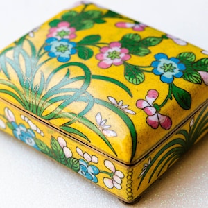 May include: A rectangular yellow metal box featuring a vibrant floral pattern with blue, pink, and green enamel details. The lid and sides are decorated with painted flowers and long green leaves, finished with a metallic trim and small feet.
