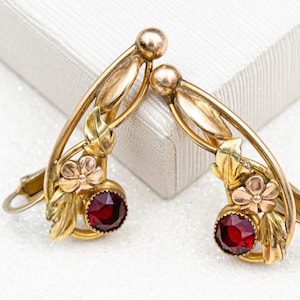 May include: A pair of antique gold-tone earrings with a floral and leaf design. Each earring features a large, round, deep red gemstone. The earrings have a curved shape with a clip-on style closure.