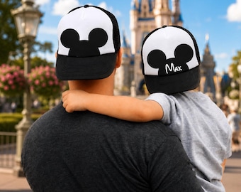 Puede incluir: Dos gorras tipo camionero con malla negra en la parte posterior y paneles frontales blancos con una silueta de orejas de ratón negra. Una gorra es lisa, mientras que la otra tiene el nombre Max escrito en blanco sobre el gráfico frontal.