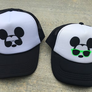 Puede incluir: Dos gorras de camionero blancas y negras con un Mickey Mouse negro con gafas de sol. Una gorra tiene gafas de sol verdes y la otra tiene gafas de sol negras.