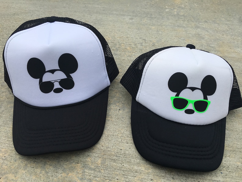 Puede incluir: Dos gorras de camionero blancas y negras con un Mickey Mouse negro con gafas de sol. Una gorra tiene gafas de sol verdes y la otra tiene gafas de sol negras.
