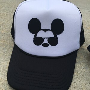 Puede incluir: Una gorra de camionero blanca y negra con un Mickey Mouse negro con gafas de sol en la parte delantera.