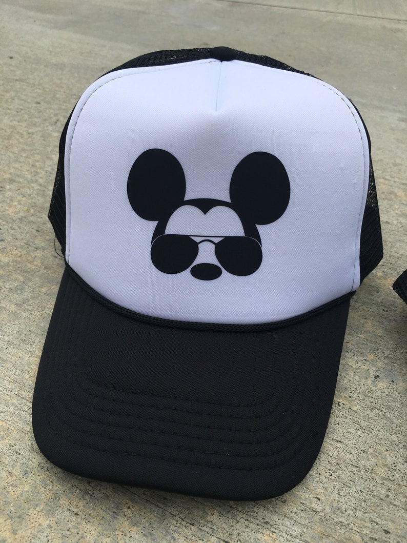 Puede incluir: Una gorra de camionero blanca y negra con un Mickey Mouse negro con gafas de sol en la parte delantera.