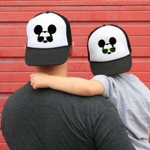 Puede incluir: Un gorro de camionero blanco y negro con un dise&ntilde;o de Mickey Mouse con gafas de sol. El gorro lo lleva una persona que sostiene a un ni&ntilde;o que lleva un gorro similar.