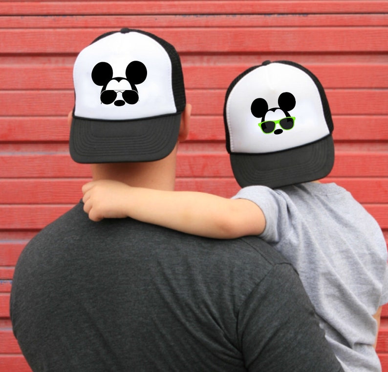 Puede incluir: Un gorro de camionero blanco y negro con un dise&ntilde;o de Mickey Mouse con gafas de sol. El gorro lo lleva una persona que sostiene a un ni&ntilde;o que lleva un gorro similar.