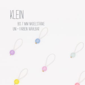 Könnte beinhalten: Eine Sammlung kleiner, farbiger Perlen an weißen elastischen Schlaufen. Die Perlen sind in verschiedenen Pastellfarben wie Blau, Rosa, Gelb und Lila gehalten. Das Wort "KLEIN" steht oben, darunter Text.