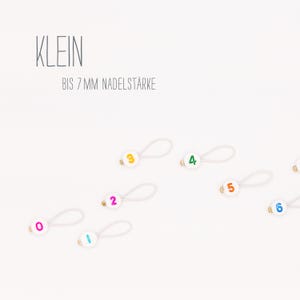 Könnte beinhalten: Weiße Kunststoff-Maschenmarkierer mit bunten Zahlen von 0 bis 6 auf weißem Hintergrund. Der Text "KLEIN BIS 7 MM NADELSTÄRKE" befindet sich oben im Bild.