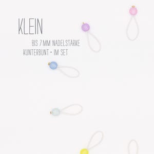 Könnte beinhalten: Ein Set aus fünf bunten, kleinen, runden Perlen mit goldenen Akzenten, jede an einer weißen elastischen Schlaufe befestigt. Die Perlen sind hellblau, helllila, hellgrün, gelb und rosa. Der Text "KLEIN" und "BIS 7 MM NADELSTÄRKE KUNTTERBUNT - IM SET" ist im Bild sichtbar.