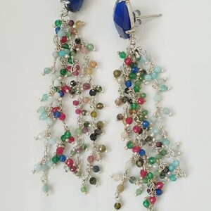 May include: A pair of silver drop earrings featuring a large, faceted, deep blue stone at the top. Below each stone, multiple silver chains hang down, adorned with small, multi-coloured, faceted gemstone beads in shades of red, green, and blue.