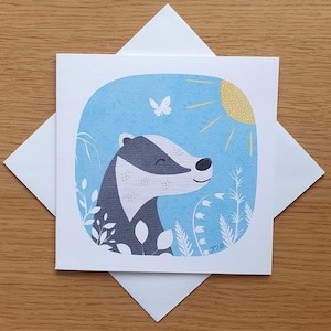 May include: A greeting card featuring a cartoon badger with a white and grey patterned body, looking up at a yellow sun with a butterfly flying above. The background is a light blue watercolour wash with white flowers and leaves.