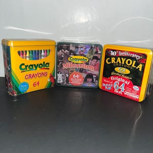 Könnte beinhalten: Drei rechteckige Metalldosen mit Crayola-Wachsmalstiften. Die linke Dose ist gelb mit einem Sichtfenster für Stifte und dem Text Crayola Crayons 64. Die mittlere Dose zeigt eine Kinder-Collage und den Text Crayola Millennium. Die rechte Dose ist gelb-schwarz mit dem Text 40th Anniversary Crayola Crayons 64 Box Limited Edition.