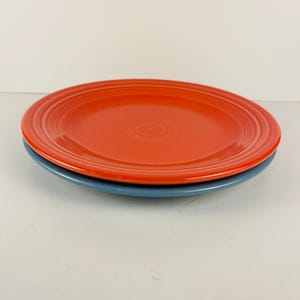 May include: A stack of two round ceramic dinner plates. The top plate is a vibrant orange color, while the bottom plate is a muted slate blue. Both plates feature a series of concentric circular ridges embossed along the outer rim.