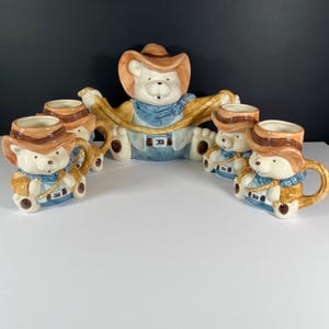 Puede incluir: Un juego de té de oso vaquero de cerámica. El juego incluye una tetera grande con forma de oso y cuatro tazas a juego. La tetera y las tazas son blancas con sombreros de vaquero marrones, pañuelos azules y detalles en color canela. Las tazas tienen asas curvas.
