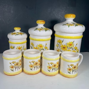 Op de afbeelding: Een vintage keramische keukenkanisterset en mokken. De set bevat drie canisters met deksels en drie mokken. De canisters en mokken zijn wit met gele madeliefjes bloemmotieven en gele randen.