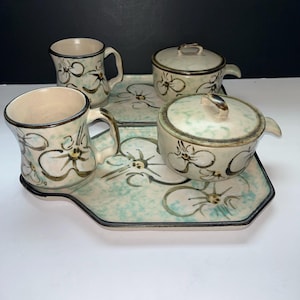 Può includere: Un servizio da tè in ceramica con un vassoio, due tazze e due contenitori con coperchio. Il set presenta una combinazione di colori verde chiaro e beige con un motivo floreale marrone e bianco. Il vassoio e i contenitori hanno un bordo nero.
