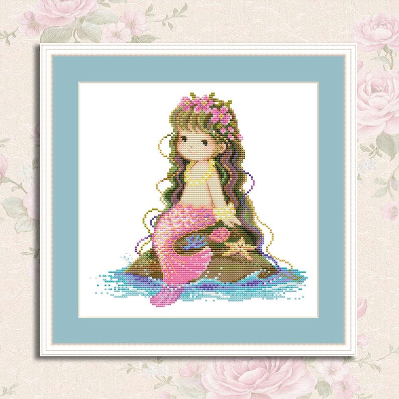 May include: A cross-stitch pattern featuring a mermaid with pink and purple scales, sitting on a rock with a flower crown. The mermaid has long brown hair and is surrounded by blue and green water.