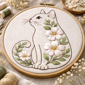 May include: Embroidered cat design in a wooden hoop. The cat is white with brown outline, adorned with white flowers and green leaves. The embroidery is set against a light beige fabric, creating a charming, handmade aesthetic.