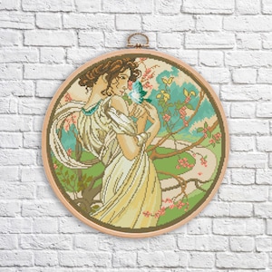 May include: A cross-stitch embroidery pattern of a woman in a white dress with a blue bird in her hand. The woman is standing in a garden with flowers and trees. The embroidery is framed in a round wooden hoop.