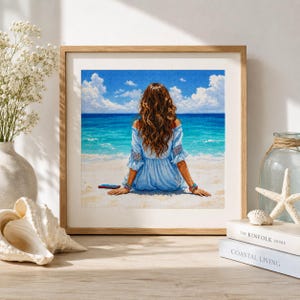 Puede incluir: Una obra de arte en punto de cruz en marco cuadrado que muestra a una persona de largo cabello castaño vista desde atrás, con un vestido azul claro de hombros descubiertos, sentada en una playa de arena frente a un océano turquesa.