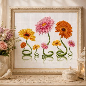 May include: A framed cross-stitch embroidery featuring a collection of stylized gerbera daisies in shades of pink, orange, and yellow. The flowers have long, curved green stems and are arranged on a white fabric background within a decorative frame.