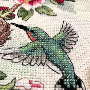 May include: A cross-stitch embroidery piece on white fabric featuring a hummingbird in flight with green and iridescent thread. The design includes pink and white flowers with green leaves and a brown nest, all rendered in detailed, textured cross-stitch.