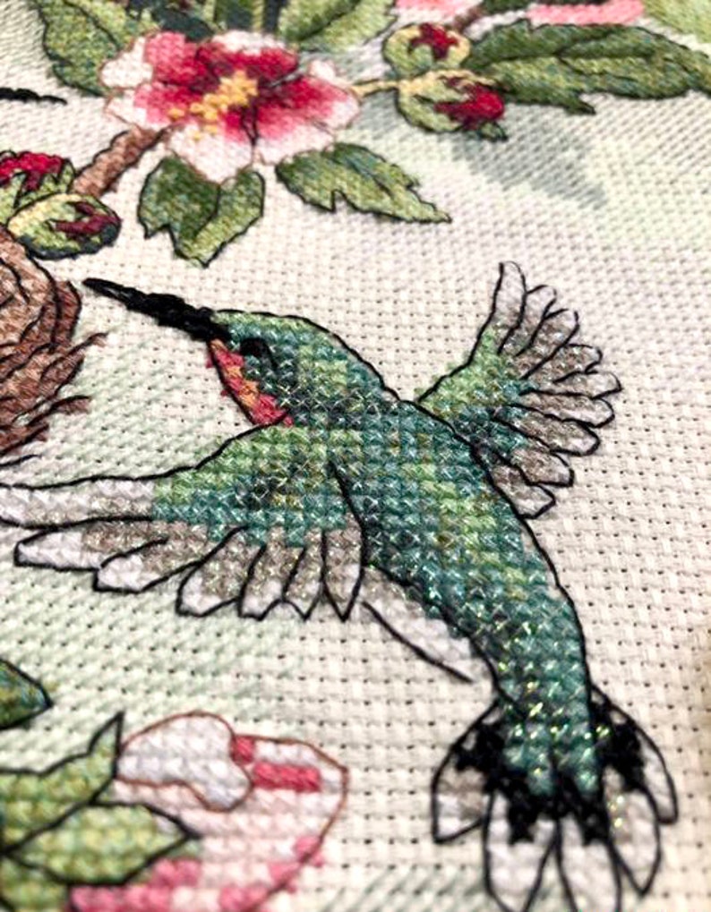 May include: A cross-stitch embroidery piece on white fabric featuring a hummingbird in flight with green and iridescent thread. The design includes pink and white flowers with green leaves and a brown nest, all rendered in detailed, textured cross-stitch.
