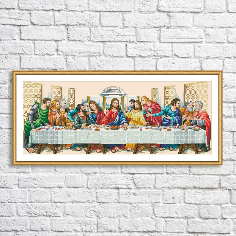 May include: A cross-stitch embroidery pattern depicting The Last Supper, featuring thirteen figures seated at a long table covered with a white patterned cloth, set within a gold-toned rectangular frame against a white brick wall background.