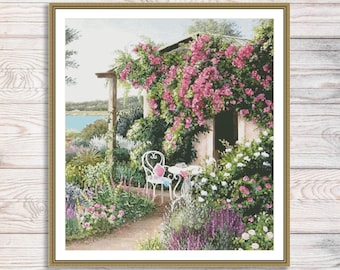 May include: A white wrought iron table and chair set with a pink cushion on the chair, set in a garden with a white cottage in the background. The cottage is covered in pink climbing roses.