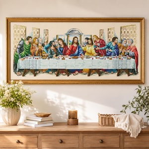 May include: A framed cross-stitch embroidery depicting The Last Supper, featuring Jesus and the twelve disciples seated at a long table covered with a white cloth, set against a light background within a decorative gold-colored rectangular frame.