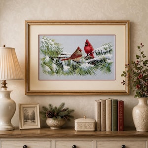 May include: A framed cross-stitch embroidery featuring two red cardinals perched on snow-covered pine branches against a light blue background. The artwork is enclosed in a gold-toned frame with a cream-coloured mat and a thin red inner border.