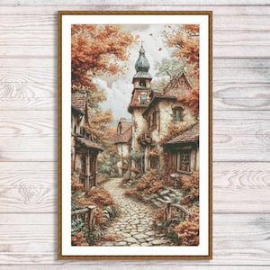 May include: A framed print of a European village scene. The artwork depicts a cobblestone path winding through buildings with red-tiled roofs and a church tower. Autumn foliage in shades of orange and brown frames the scene, creating a warm, inviting atmosphere.