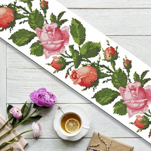 Puede incluir: Un patrón de bordado de punto de cruz blanco con un borde de rosas rosas y naranjas con hojas verdes. El bordado está sobre un fondo de tela blanca.
