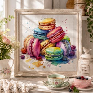 May include: A framed cross-stitch embroidery featuring a colorful stack of macarons in shades of yellow, blue, pink, purple, and green, accented with scattered berries and watercolor-style paint splatters on a white fabric background.