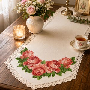 Könnte beinhalten: Ein beigefarbener, rechteckiger Tischläufer mit einem Kreuzstichmuster aus rosa Rosen und grünen Blättern am spitz zulaufenden Ende. Der Tischläufer ist an den Rändern mit einer feinen weißen Spitzenborte verziert.