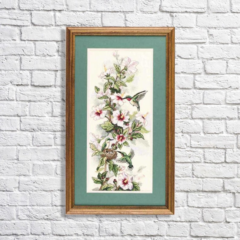 May include: A framed cross-stitch embroidery featuring two hummingbirds and a nest among white and pink hibiscus flowers. The artwork is encased in a wooden frame with a green mat border, showcasing detailed needlework of birds and floral stems.