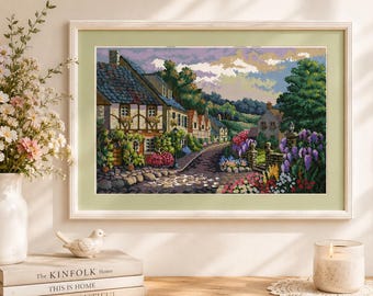 May include: A framed cross-stitch embroidery depicting a European village scene with stone houses, a cobblestone path, and vibrant flower gardens. The artwork features a purple and white sky, green trees, and colorful floral clusters in the foreground.