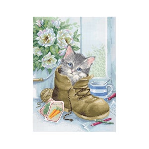 May include: A gray and white kitten with blue eyes sits inside a brown leather boot. The boot is on a table with a cup, a ball of yarn, and seed packets.
