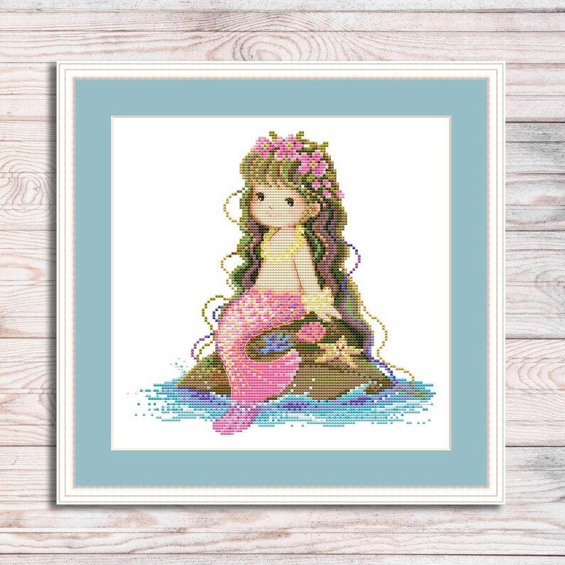 May include: Cross stitch pattern of a mermaid with pink tail and a flower crown sitting on a rock with blue water behind her.