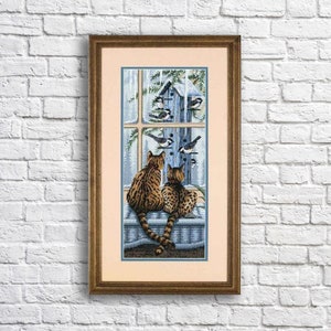 May include: A framed cross-stitch artwork depicts two cats with brown and black patterned fur, gazing out a window at birds and a birdhouse. The artwork is set against a light blue background and framed in a brown wooden frame.