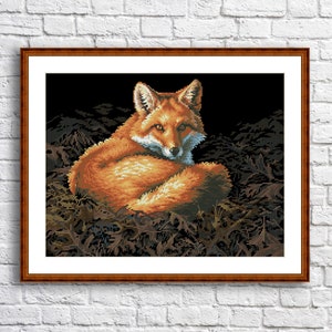 May include: A framed cross-stitch of a red fox resting in a bed of brown leaves. The fox has orange and white fur, with a dark background. The artwork is in a brown wooden frame and hangs on a white brick wall.