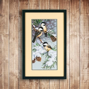 May include: Cross-stitch picture of three chickadees on snow-covered pine branches. The birds have black caps, white cheeks, and tan bodies. The artwork is framed in a black frame with a cream-coloured mount, set against a wooden background.