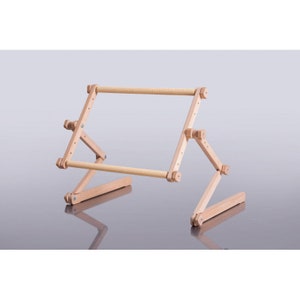 May include: A light-coloured wooden embroidery hoop stand. The stand is adjustable, with a rectangular frame held by hinged wooden supports. The frame has a textured, cylindrical bar on each side for holding fabric. The stand is designed to sit on a flat surface.
