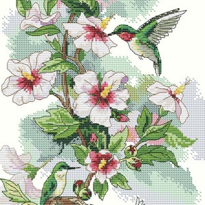 May include: A cross-stitch pattern featuring three hummingbirds with green and red plumage interacting with a flowering hibiscus plant. One bird hovers near a bloom, another sits in a nest with eggs, and a third flies near the flowers.