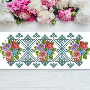 Könnte beinhalten: Ein Kreuzstich-Stickmuster mit einem floralen Rand mit rosa, roten und violetten Blumen auf weißem Hintergrund. Das Design enthält ein geometrisches Muster aus blauen und grünen Quadraten.