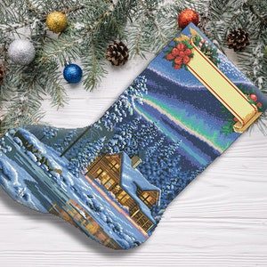 May include: A blue and white Christmas stocking featuring a snowy cabin scene and a blank tag. The stocking has a white border and a red and green holly design at the top.