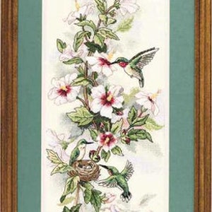 May include: A framed cross-stitch embroidery featuring three hummingbirds and a nest among white hibiscus flowers with pink centres. The artwork is set against a cream background, enclosed in a green mat and a rectangular wooden frame.