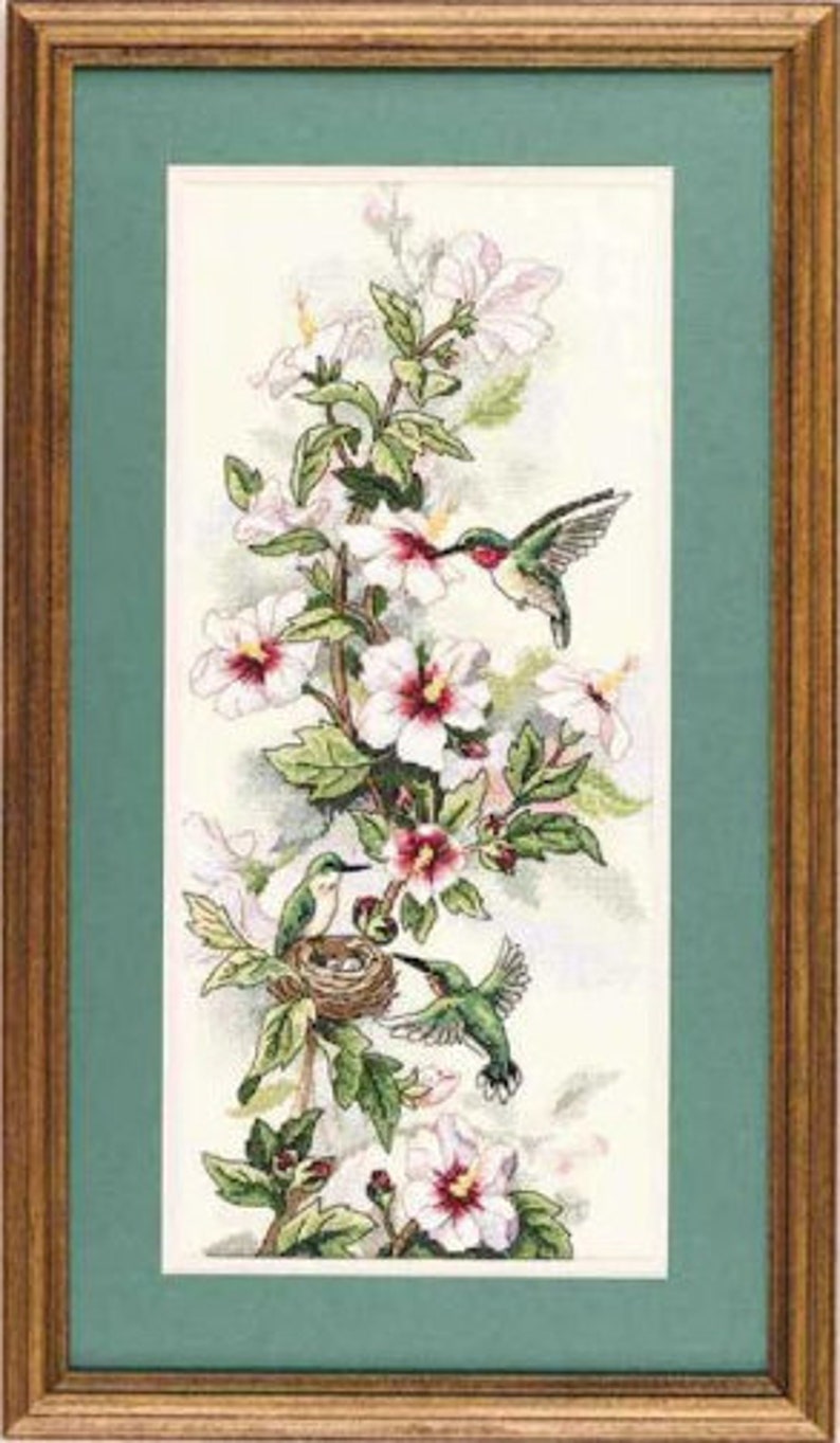 May include: A framed cross-stitch embroidery featuring three hummingbirds and a nest among white hibiscus flowers with pink centres. The artwork is set against a cream background, enclosed in a green mat and a rectangular wooden frame.